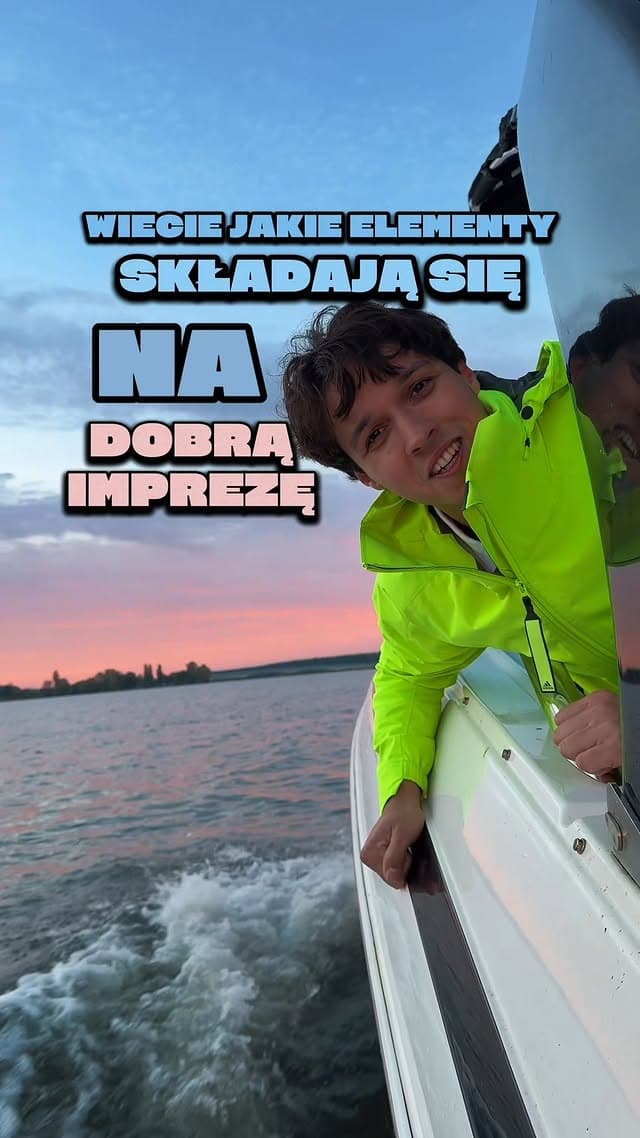 Członek załogi OtoRejs w żółtej kurtce wychyla się z jachtu płynącego o zachodzie słońca